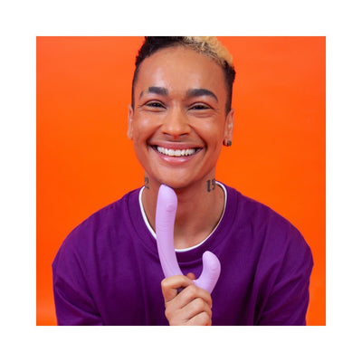 Smiling person in purple shirt holding a lavender curved dual-ended intimate toy against an orange background.