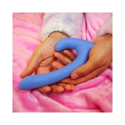 Hands holding a curved blue silicone dual-ended intimate toy on a soft pink blanket background.