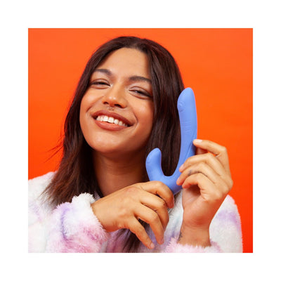 Smiling woman holding a blue silicone dual-stimulation adult toy against an orange background.