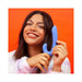 Smiling woman holding a blue silicone dual-stimulation adult toy against an orange background.