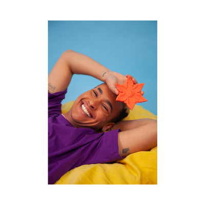 Smiling person lying on yellow bedding holding a rose-colored star-shaped textured companion toy against a blue background.