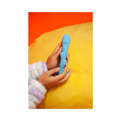 Hands holding a teal silicone toy with playful faces, set against a bright yellow and orange background.