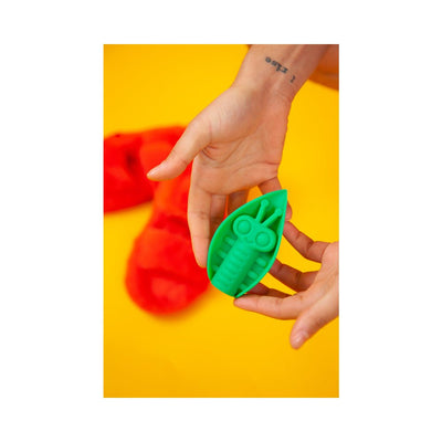 Hands holding a green leaf-shaped silicone vibrator with textured insect design against a yellow background.