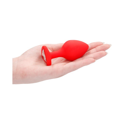 Hand holding a large red silicone butt plug with a clear diamond-shaped gem at the flared base.