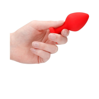 Hand holding a large red silicone butt plug with a tapered shape and flared base against a white background.