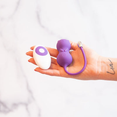 Hand holding purple cat-shaped kegel ball vibrator and white remote control on marble background