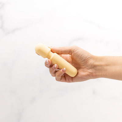 Hand holding a compact, textured pale yellow Emojibator Tiny Wand vibrator against a white background.