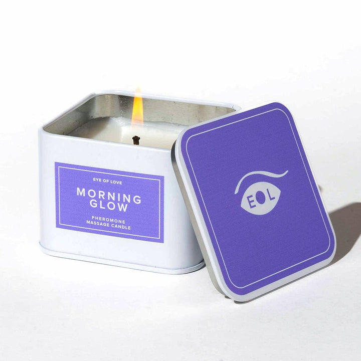 Lit Eye of Love Morning Glow pheromone massage candle in a white square tin with a purple lid beside it.