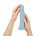 Hands holding a light blue realistic textured silicone vibrator with a rounded tip and control buttons near the base.