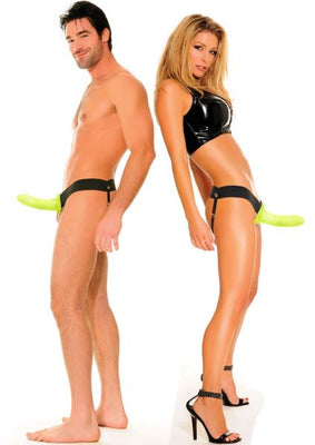 Man and woman wearing black harnesses with neon green hollow strap-ons, posing back to back on white background.