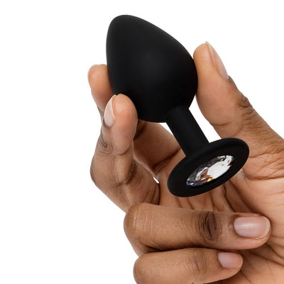 Hand holding a black silicone butt plug with a clear crystal base detail on a white background.