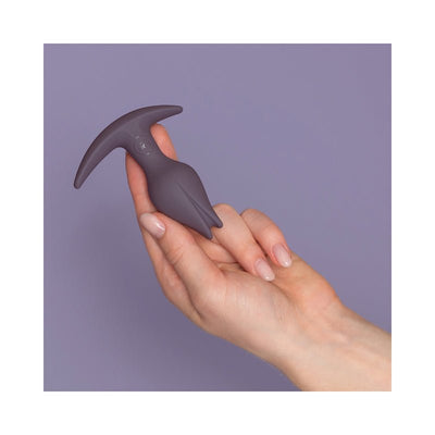 Hand holding a small, dark purple silicone butt plug with an asymmetrical tulip-shaped tip and a T-shaped base against a lavender background.