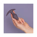 Hand holding a small, dark purple silicone butt plug with an asymmetrical tulip-shaped tip and a T-shaped base against a lavender background.