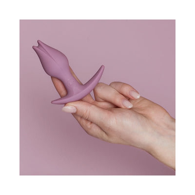 Hand holding a small mauve silicone butt plug with a tulip-shaped tip and asymmetrical base against a matching mauve background.