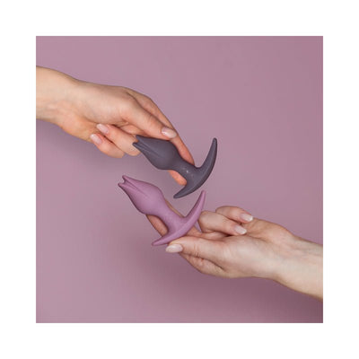 Two tulip-shaped silicone butt plugs in mauve and dark gray held by hands against a soft purple background.