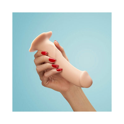 Hand holding a vanilla-colored silicone dildo with a pronounced head and suction base against a light blue background.