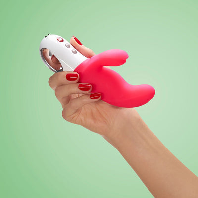 Hand holding a pink and white dual-action vibrator with ergonomic handle and control buttons on a green background.