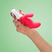 Hand holding a pink and white dual-action vibrator with ergonomic handle and control buttons on a green background.