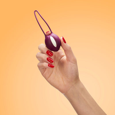 Hand holding a white and grape-colored Fun Factory Smartball Uno pelvic floor trainer against a peach background.