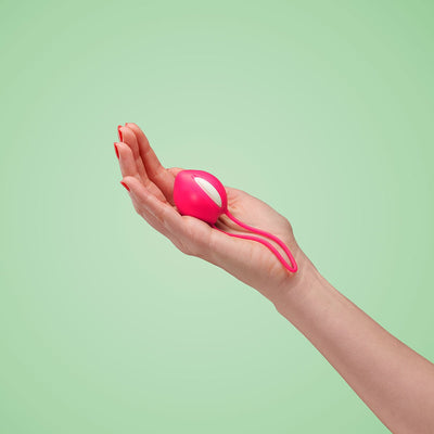 Hand holding a pink and white Fun Factory Smartballs Uno Kegel exercise ball against a light green background.