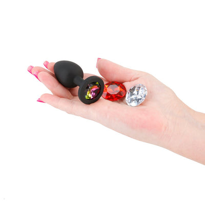 Hand holding a black silicone plug with three interchangeable colorful gem attachments in pink, red, and clear.