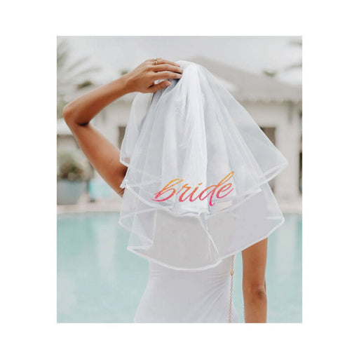 Bride wearing a double-layered white tulle veil with rose-gold "bride" lettering at the back by a poolside.