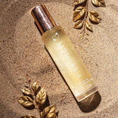 Highonlove Dry Body Oil Spray bottle with golden cap on textured sandy surface with decorative gold leaves.
