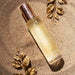 Highonlove Dry Body Oil Spray bottle with golden cap on textured sandy surface with decorative gold leaves.