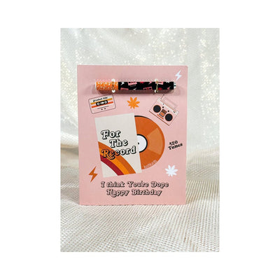 Pink birthday card with retro music design and attached colorful one hitter pipe accessory.