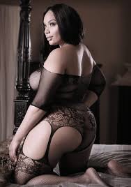 Woman wearing sheer black off-shoulder teddy net gartered bodystocking with floral lace details, posing on bed.