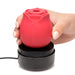 Hand holding red rose-shaped stimulator on black charging base with USB cable on white background
