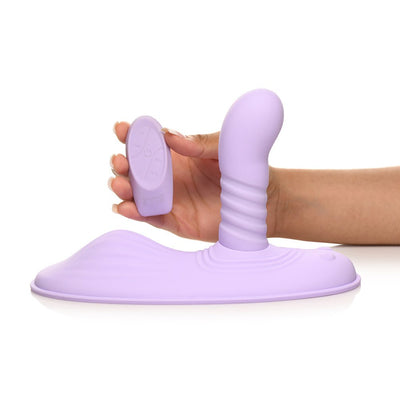 Hand holding remote control next to a purple silicone thrusting and vibrating adult toy with a curved shaft and textured base.