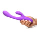 Hand holding a purple silicone rabbit vibrator with curved shaft and flexible flicking arm against a white background.