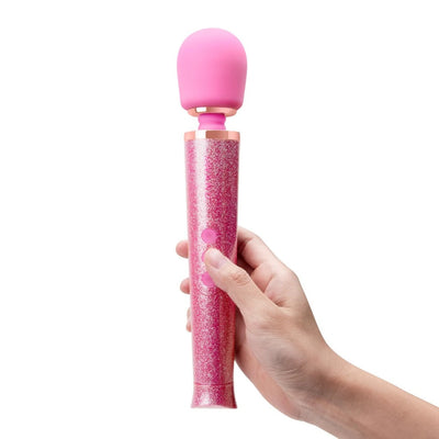 Hand holding a pink glittery Le Wand Petite massager with a rounded head and two control buttons.