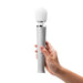 Hand holding a sleek silver Le Wand Petite massager with a white rounded head and three control buttons.