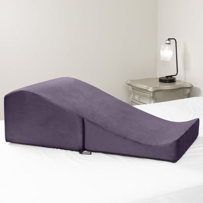 Plum-colored Liberator Flip Ramp wedge pillow on a white bed with a nightstand and lamp in the background.