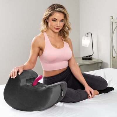 Woman seated on bed next to black Liberator Pulse Toy Mount with pink toy inserted, in a bright bedroom setting.