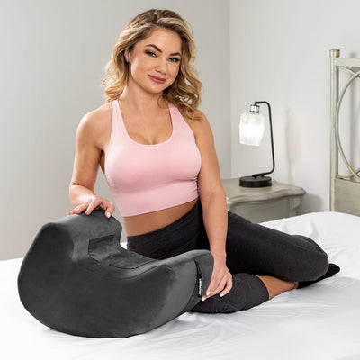 Woman in pink sports bra and black leggings sitting on bed next to a black Liberator Pulse Toy Mount with soft velvet cover.
