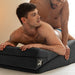 Two men using a black Liberator Wedge/Ramp Combo cushion for support during intimate moments on a bed.
