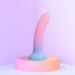 Glow-in-the-dark silicone dildo with a curved design in pastel pink and blue on a round pink pedestal.