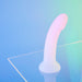 Curved pastel pink dildo with glitter finish standing on clear surface against a soft blue and green gradient background.