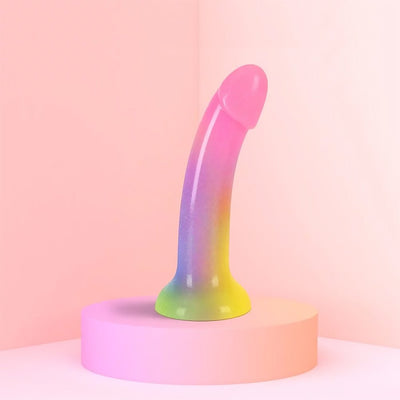 Curved rainbow-colored silicone dildo with glitter and suction cup base on a pink pedestal.