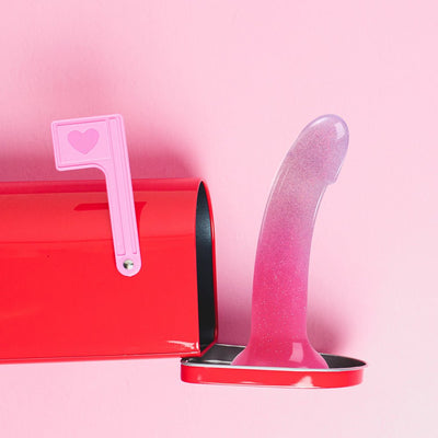 Pink and purple gradient silicone dildo with multicolor star glitter standing inside a red mailbox against a pink background.