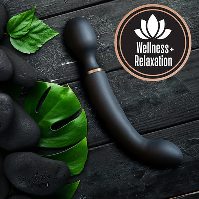 Black dual-ended massager with curved design on dark wood background, surrounded by green leaves and black stones, wellness and relaxation theme.