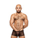 Muscular man wearing black sheer mini shorts with a small heart pattern and a contoured pouch, posing against white background.