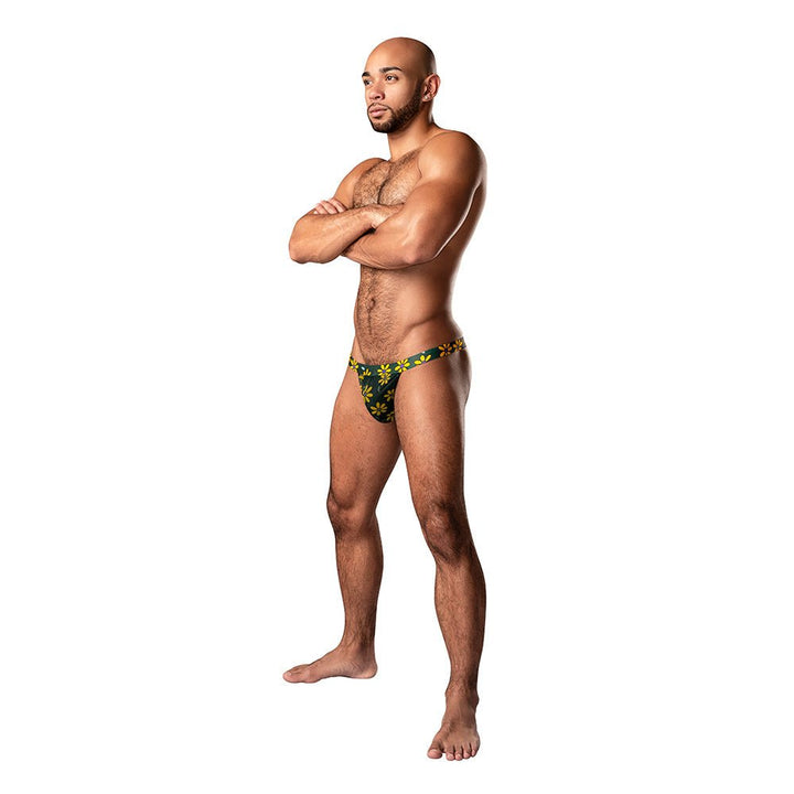 Male model wearing Male Power Petal Power Micro Thong with yellow daisy print on dark green sheer mesh fabric.