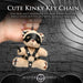 Hand holding a small peach teddy bear keychain wearing a black faux leather bondage harness, eye mask, and holding a mini flogger.