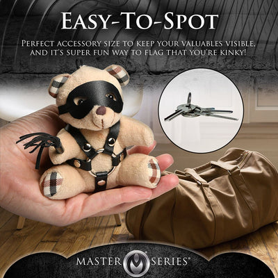 Hand holding a small peach teddy bear keychain wearing a black faux leather harness, eye mask, and holding a mini flogger next to a brown bag.