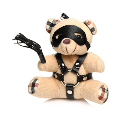 Small beige teddy bear keychain wearing black faux leather harness and eye mask, holding a mini flogger with plaid ear and paw accents.