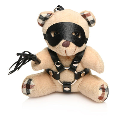 Small plush teddy bear keychain with plaid ear and paw details, wearing a black faux leather harness, eye mask, and holding a mini flogger.